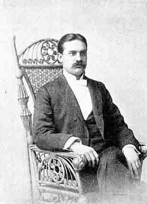 White bow tie, dark three-piece double-breasted suit; seated in ornate bentwood and cane chair.